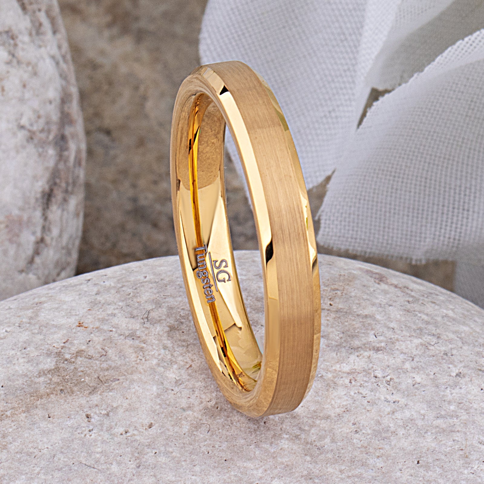 Yellow gold tungsten wedding ring with brushed finish and beveled edges, shown on a stone surface.